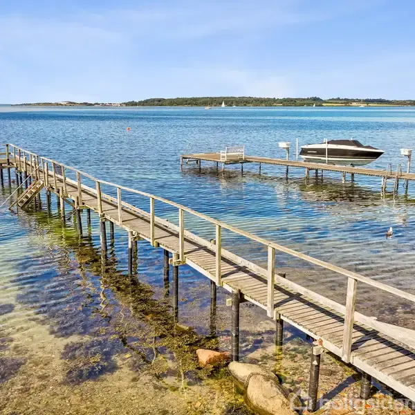 En træbro strækker sig ud over klart vand fyldt med tang. En mindre motorbåd og en vandscooter ligger fortøjet ved en parallel bro. I baggrunden ses en skovklædt kystlinje.