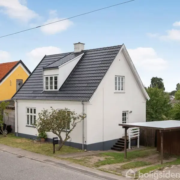 Hvidt hus med gråt tag står ved siden af et gult hus. Der er en carport ved siden af huset, omgivet af græsplæne og træer, under en blå himmel.