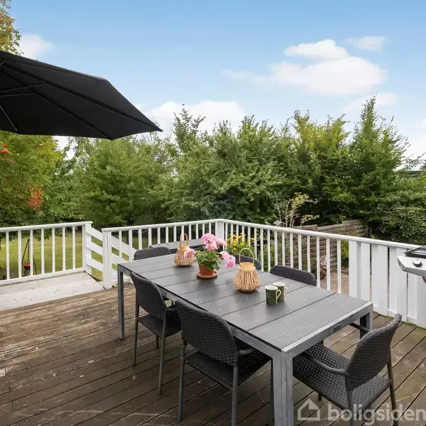 Terrassebord står dækket med blomsterdekorationer og seks stole, under en stor parasol. I nærheden er en grill placeret. Omgivet af træer og grønne buske i en have.