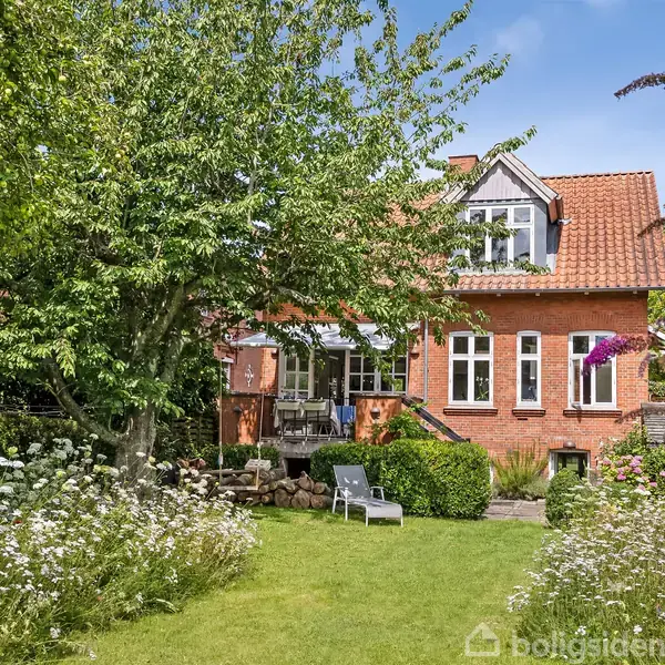Hus i mursten med rødt tegltag står i frodig have. To liggestole er placeret på græsplænen omgivet af vilde blomster og træer, der skaber en afslappet, naturlig atmosfære.