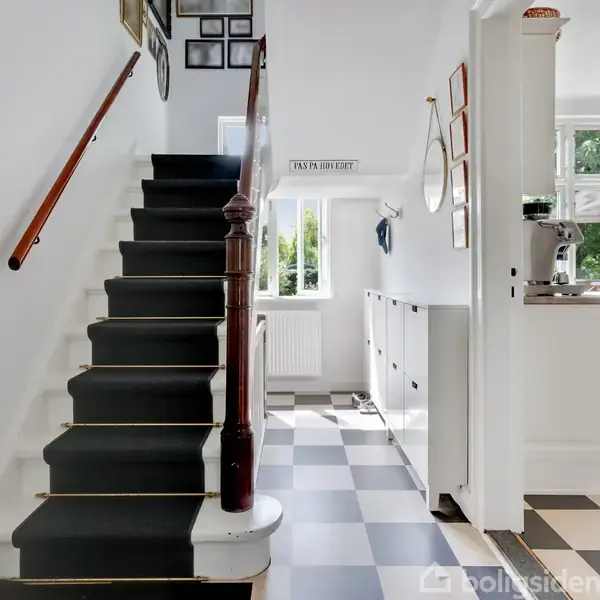 En trappe med trægelænder fører opad i en lys indgang med rutet gulv. En dør til højre åbner ind til et rum med køkkenudstyr. Tekst: "FALKA ER EN LIAR."