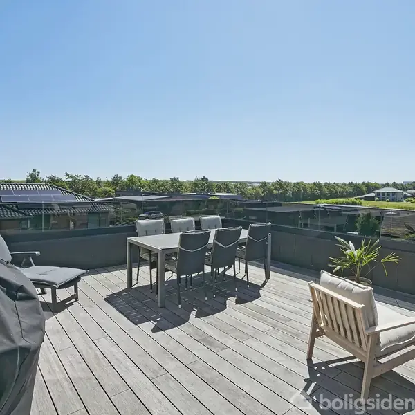 En rummelig terrasse med havemøbler og grill på trædæk, omgivet af beboelsesbygninger og grønne områder under en klar blå himmel.