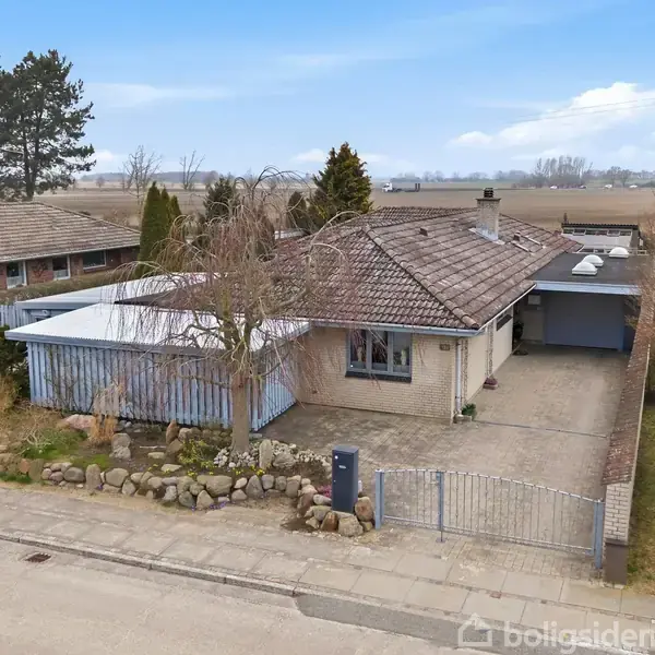 Et enfamiliehus med fladt tag står i et villakvarter med en indkørsel og carport. Det omkranses af træer og en lav murstensmur. Baggrunden viser åbent land.