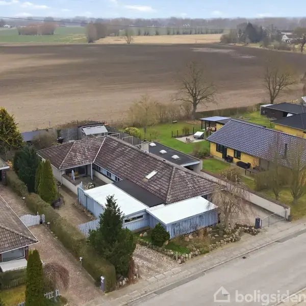 Villa med skråtag står stille langs en stille vej. Omkranset af andre huse, har den en have med marker i baggrunden. Det er en landskabelig landsbyatmosfære.