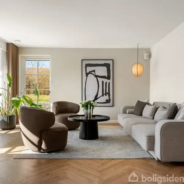 Stue med grå sofa og to lænestole omkring et rundt sofabord. Store vinduer, planter og minimalistisk kunst præger rummet, der har trægulv og en hængende lampe.