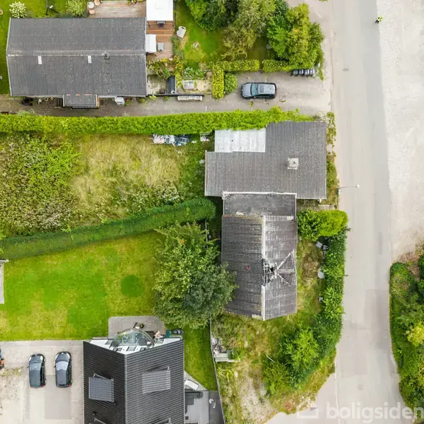 Overblik af en bolig med et delvist ødelagt tag, omgivet af grønne haver og veje. Flere huse og biler kan ses rundt omkring i et beboelsesområde.
