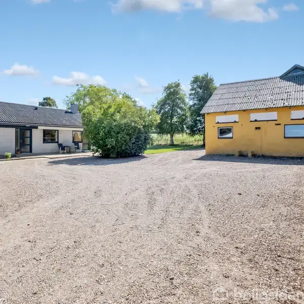 En åben gårdsplads med grus, omgivet af to bygninger; en grå bygning til venstre og en gul bygning til højre. I midten står en stor busk.