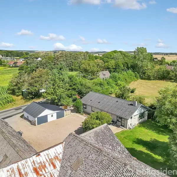 Hus set oppefra omgivet af grønne træer og marker; ejendommen har tilhørende garage og grusbelagt gårdsplads. Landlig beliggenhed med åbne marker i baggrunden.