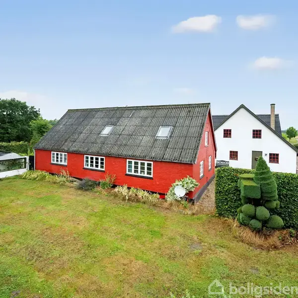 Rød bygning med skråt tag står stille ved siden af en hvid bygning med gavl. Omgivet af grøn have og trimmede buske under en blå himmel.