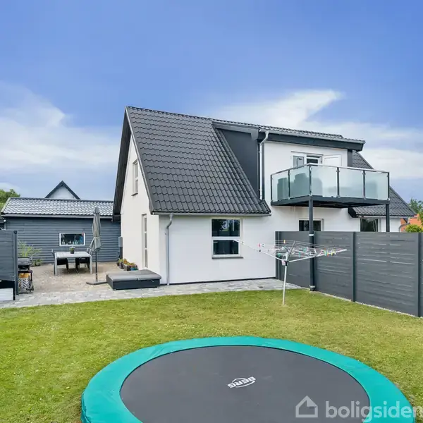 Et to-etagers hus med balkon, omgivet af en lukket have. Foran i haven er en nedgravet trampolin, og til venstre ses et flisebelagt terrasseområde med grill samt havemøbler.