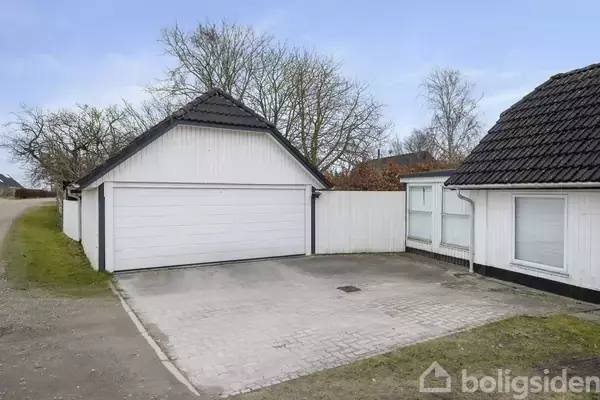 Hvid garage med sort tag står ved siden af en lille gårdsplads, flankeret af et hus. Træer uden blade ses i baggrunden på den klare dag.