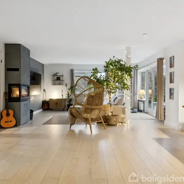 Stue med pejs til venstre og trægulv. En kurvestol står midt i rummet, omgivet af grønne planter. Store vinduer giver udsigt til haven udenfor.