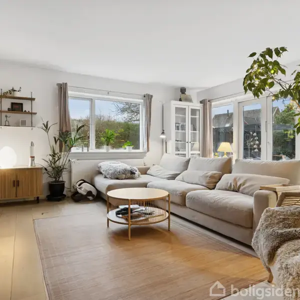 Stue med en stor, beige sofa foran vinduesparti, omgivet af grønne planter. Rundt sofabord står i midten, mens en rattan lænestol med tæppe står til højre. Tv på venstre væg.