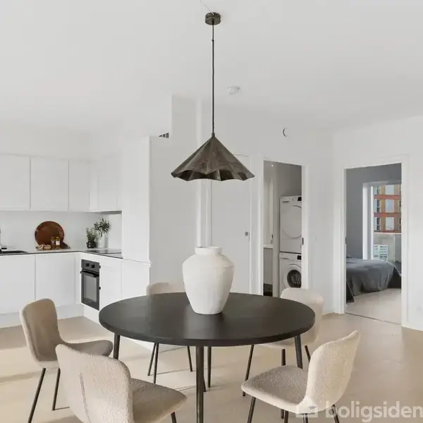 Rundt spisebord med fem stole står i et lyst, moderne køkken-alrum. En stor vase pryder bordet. Baggrund: Hvidt køkken med indbygget ovn, og to døråbninger fører til andre rum.