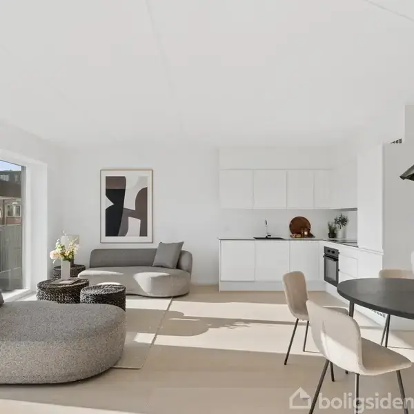 Stue med moderne indretning; gråt, kurvet møbel og sofabord ved vindue. Åben hvidt køkken i baggrunden. Rundt spisebord med stålgrå lampe. Kunstværk på væggen og lysindfald fra venstre.