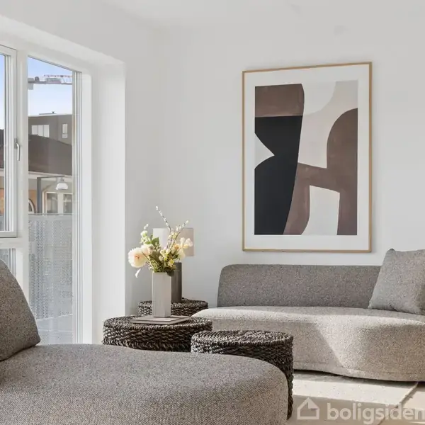 Grå sofaer står i en lys stue ved siden af et rundt sofabord og en vase med blomster. En abstrakt kunstplakat hænger på væggen ved siden af et stort vindue.