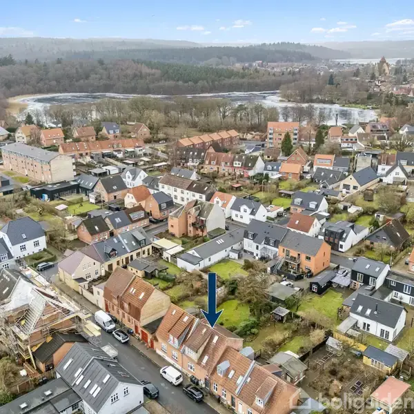 En by set oppefra med tætliggende huse med røde tage. En blå pil peger mod et specifikt hus. Landskabet inkluderer bakker og træer i baggrunden.