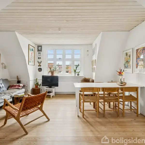 Sofa står ved væggen, omgivet af malerier og planter. Et spisebord med stole er på højre side under skråloftet. Store vinduer lukker lys ind i det hyggelige rum.