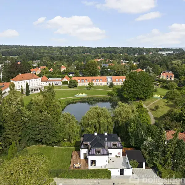 Et hvidt hus med sorttag står i forgrunden omgivet af frodig vegetation. I baggrunden ses en lille sø og flere rødtagsbygninger i en bymæssig, grøn landskabsomgivelse.