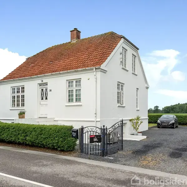 Hvidt hus med rødt tag ligger ved siden af en vej. En bil holder parkeret på grusindkørsel. Hækken omkranser haven, mens marker strækker sig i baggrunden. Ingen tekst.