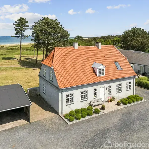 Hvidt hus med rødt tag står stille i et kystområde, omgivet af grusvej, carport, og græsklædt landskab med træer og havudsigt i baggrunden.