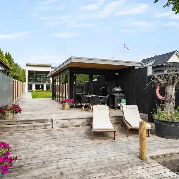Liggestole står stille på en træterrasse ved siden af et moderne udendørs køkken med grill. Omkring er der blomsterkrukker, en sortmalet væg og en velholdt have i baggrunden.