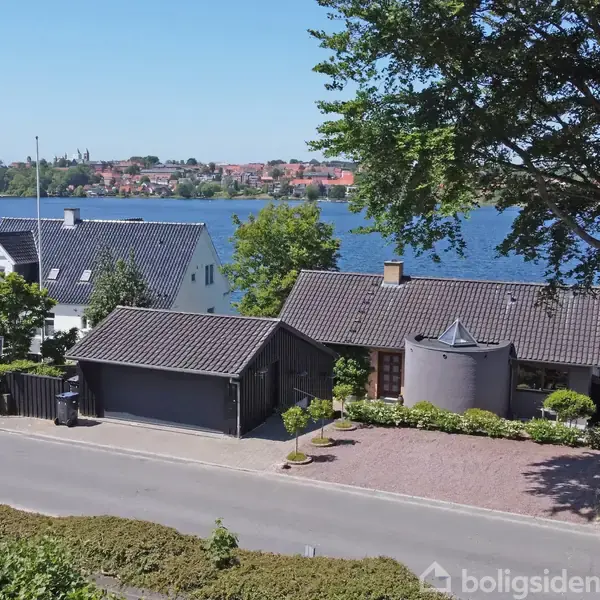 Hus står ved en sø med en anlagt have og en stor træterrasse. Der er en garage ved siden af og vejen foran huset. Skoven og byen kan ses i baggrunden.