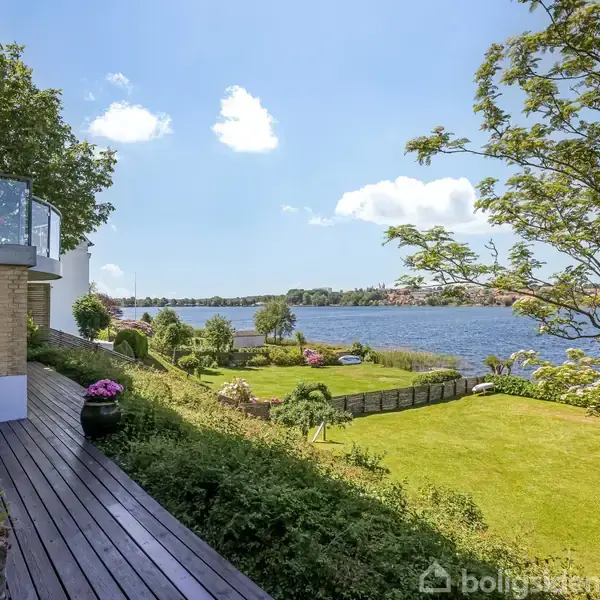 Træterrasse strækker sig langs hus, omgivet af grøn beplantning, med udsigt over en stor, velholdt græsplæne, der munder ud ved en rolig sø under en klar blå himmel.