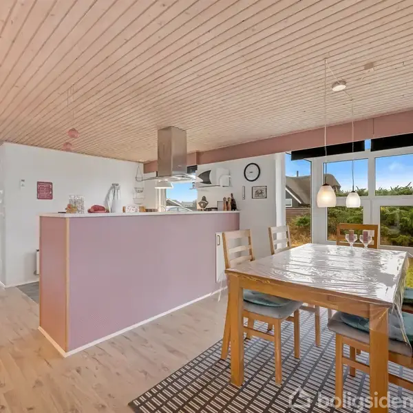 Et spisestuebord med træstole er placeret på et tæppe i et rum med træloft, store vinduer og et åbent køkkenområde med lyserøde vægge og moderne dekoration.