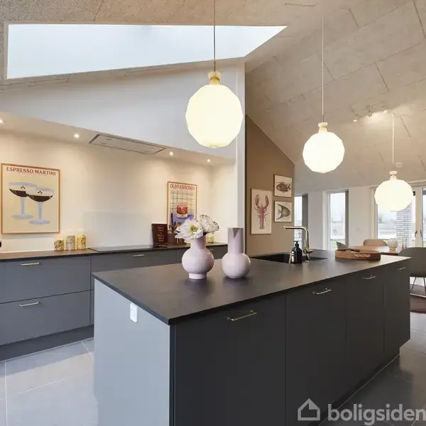 En moderne køkkenø med sorte borde står under hængende lamper. Køkkenet har store vinduer og billeder på væggene, hvilket skaber en lys og elegant atmosfære.