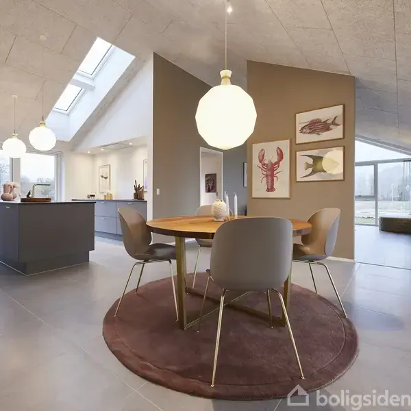 Rundt træbord med fire stole står på brun tæppe; over bordet hænger en stor lampe. I baggrunden ses åbent køkken og illustrerede billeder på væggen i moderne lyse stue.