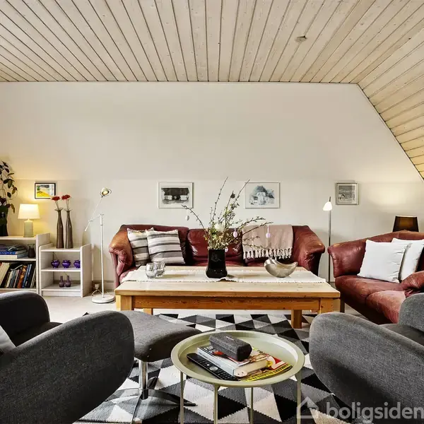 Stue med grå lænestole og røde sofaer omkring et træbord, dekoreret med bøger og vaser, på et geometrisk mønstret tæppe. Væggene har billeder, og en bogreol står i hjørnet.
