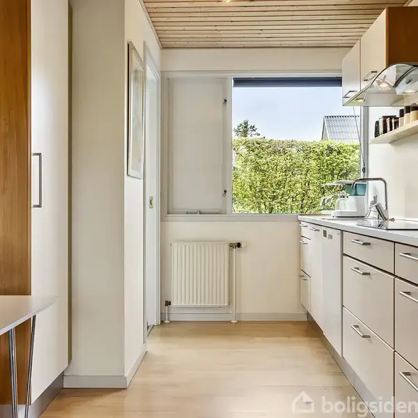 Et moderne køkken med hvide skabe og indbygget ovn. En spisebord med blomster i forgrunden. Der er et vindue med udsigt til en hæk, som giver naturligt lys.