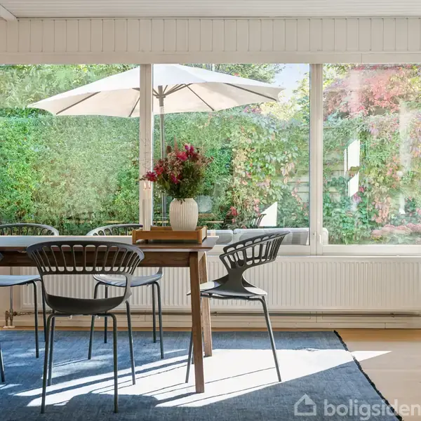 Spisebord med seks stole står på et tæppe i en lys spisestue. Kander med blomster dekorerer bordet. Store vinduer viser en have med parasol i baggrunden.