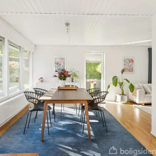 Spisebord med stole placeret på en blå måtte. Vaser med blomster står på bordet. I baggrunden er en sofa nær et vindue. Rummet er lyst med store ruder.