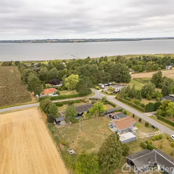 Flere huse ligger spredt i et grønt landskab nær kysten, med marker omkransende området. En vej snoer sig gennem husene, og havet er synligt i baggrunden under en overskyet himmel.