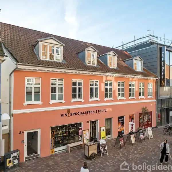 En orange bygning med skiltet "VIN SPECIALISTEN STOLTZ" ligger ved en brostensbelagt gågade. Foran butikken er der udstillede varer og gående forbipasserende i bymiljøet.