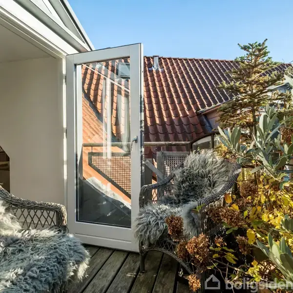 En terrasse med to kurvestole dækket af fåreskind står foran en åben glasdør. I baggrunden ses et rødt tegltag og frodige grønne planter.