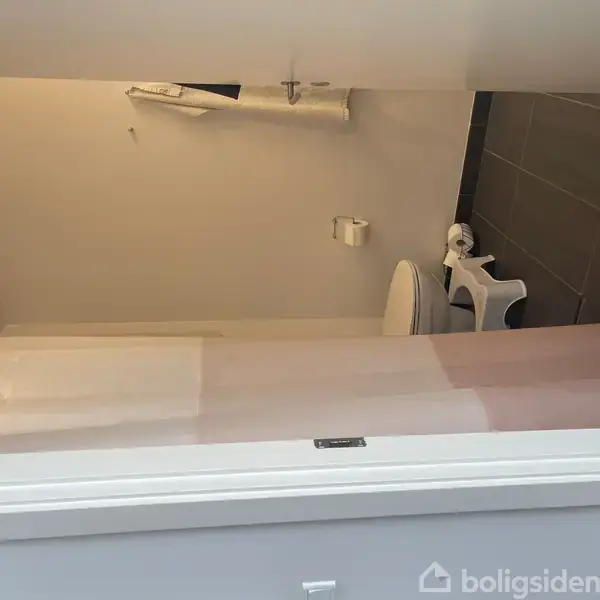 Badeværelse med toilet og bruser. Toilettet står ved væggen, og der hænger et bruseforhæng ved siden af. Der er toiletpapirholder og en lille skammel. Gulvet er flisebelagt.