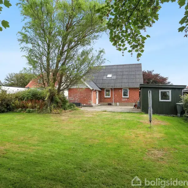 Rødt murstenshus med gråt tag står i midten af en velplejet have; til venstre et stort træ, til højre et grønt skur, og hæk omkranser græsplænen.