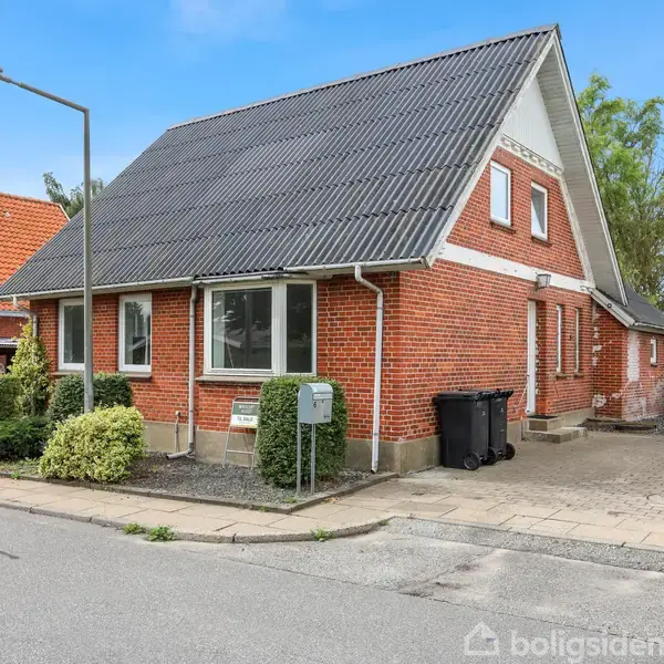 Rødt murstenshus med sort tag står stille på en stille gade med fortov. Der er nogle buske ved husets facade, og en skraldespand ved siden af indkørslen.