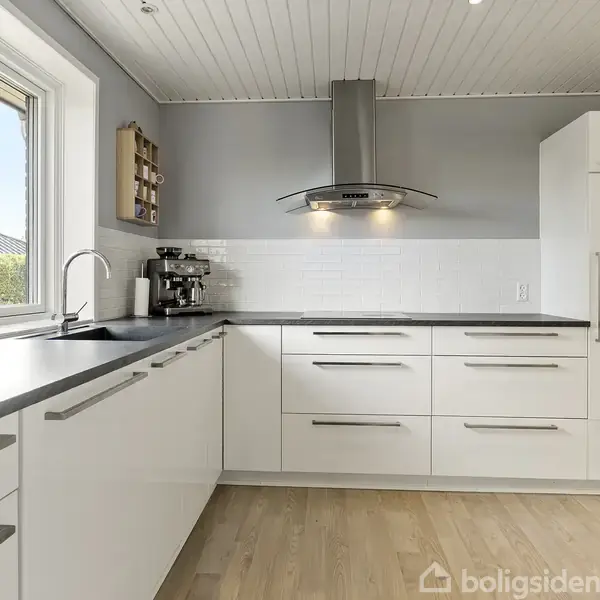 Køkkenet har hvide skabe og grå bordplade, frisket op med moderne køkkenudstyr. En emhætte hænger over en glaskeramisk kogeplade. Sollys strømmer ind fra et stort vindue til venstre.