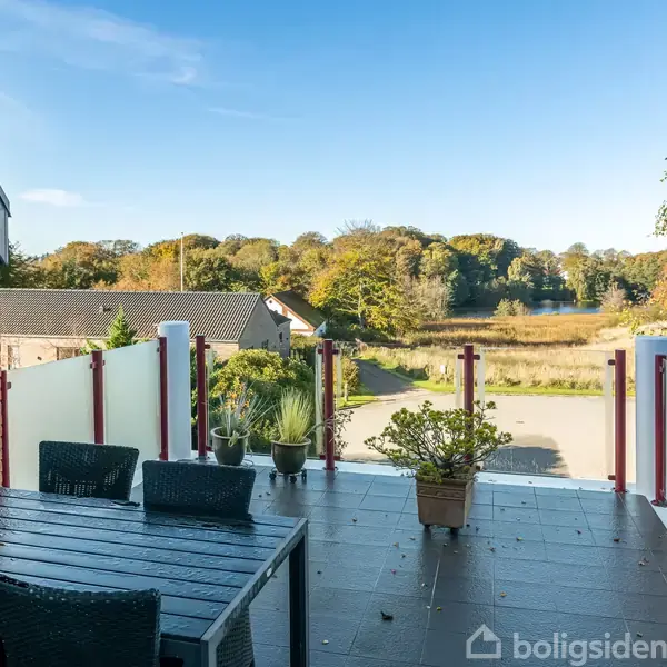 Terrasse med sorte havemøbler og planter, der står stille. Omkringliggende landskab inkluderer en vej, huse og en skov i det fjerne under en blå himmel.