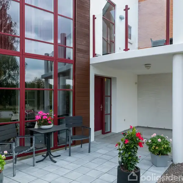 Terrasse med to stole og et bord dekoreret med blomsterkrukker. Placeret foran et hus med store vinduer og rød ramme. Området er flisebelagt og overdækket.