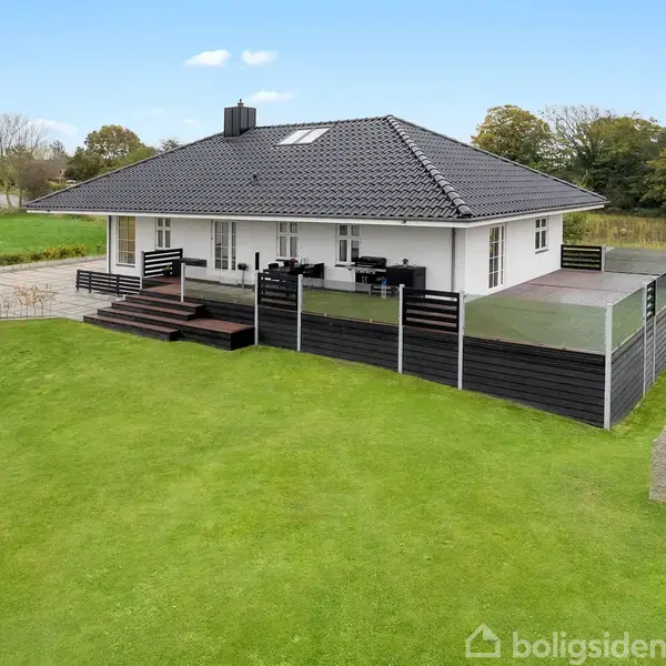 Hus med sort tag og hvid facade står stille i en grøn have. Terrassen har glasværn, og der er en stenplade på græsset.