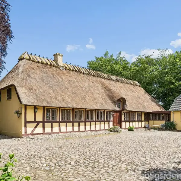 Bindingsværkshus med stråtag står i en brostensbelagt gårdsplads. Omkring huset er der grønne træer og buske under en blå himmel med enkelte skyer.