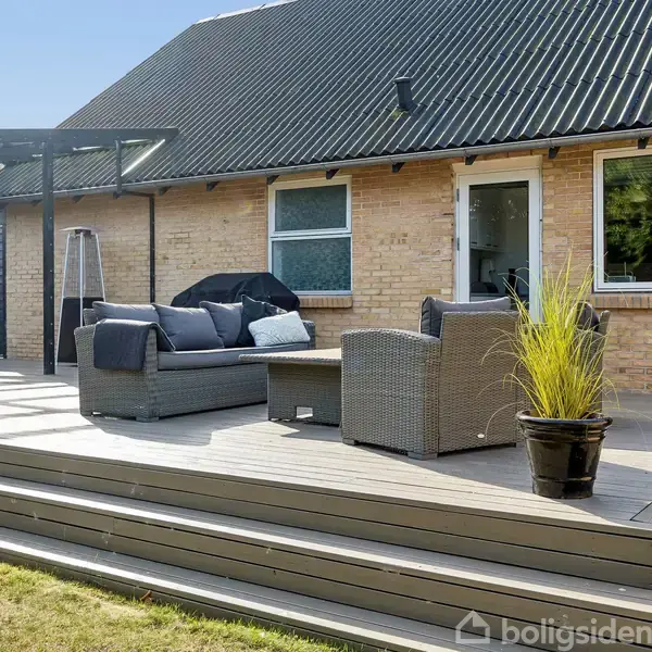 Terrasse med grå møbler og planter står foran et hus med gult murværk og sorte tagplader. Til venstre ses et udendørs spisebord under en pergola.