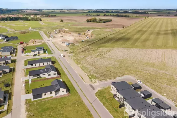 Luftfoto af et boligområde under udvikling. Moderne enfamiliehuse ligger langs en vej, omgivet af velplejede grønne plæner. Byggeplads med entreprenørmaskiner ses i baggrunden mod åbne marker.