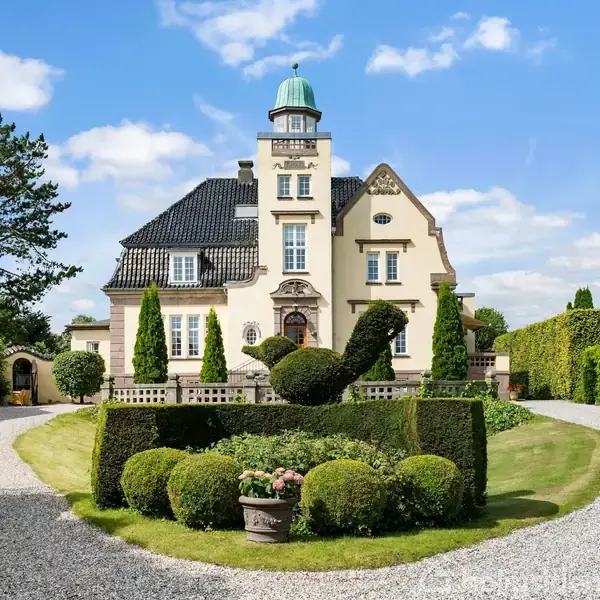 Stort hus med tårn og kuppel, omgivet af velplejet have med formklippet busk i fugleform. Beliggende på en grusvej, med grønne træer og buske omkring.