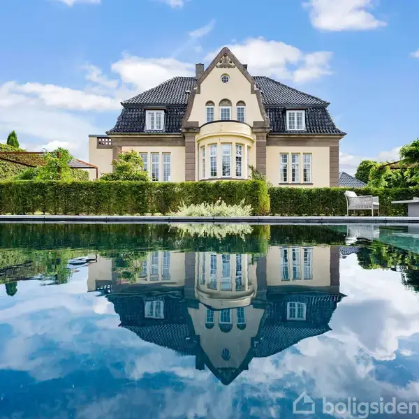 Villaen reflekteres i en rolig pool. Husets elegante facade og symmetriske vinduer fremhæves. Baggrunden er en skyfri blå himmel, omgivet af velplejet have og græs.